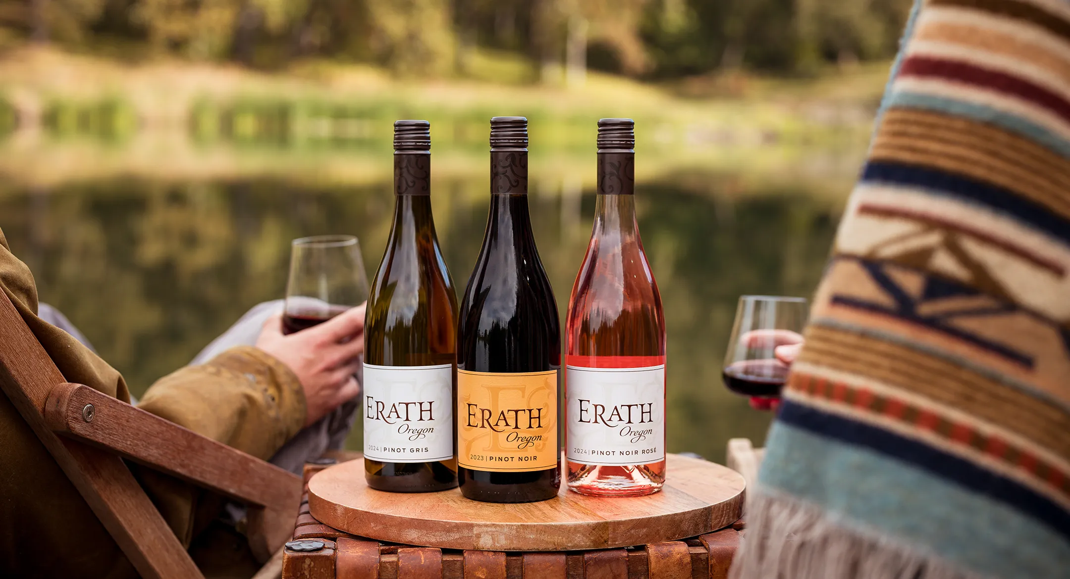 Three bottles of Erath wines on a wooden side table by a lake.