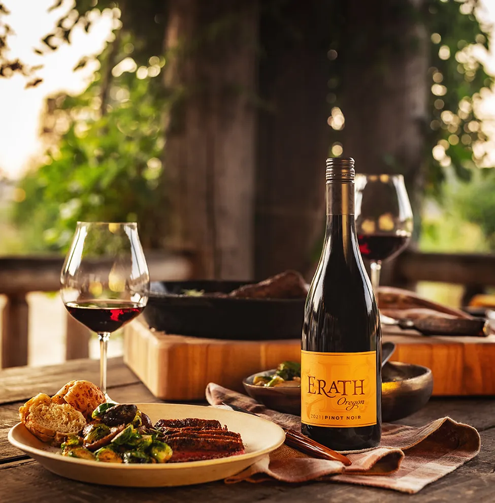 A solid wood picnic table under a large tree canopy has a plate of food and a glass of Erath wine beside the bottle of Erath pinot noir.
