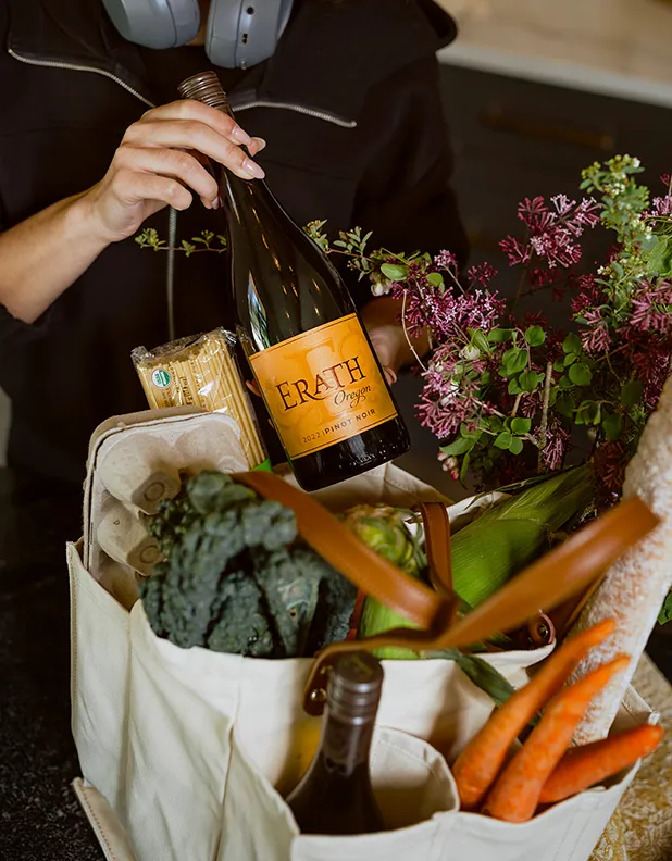 A woman lefts out a bottle of Erath pinot Noir from a shopping bag of fresh vegetables.