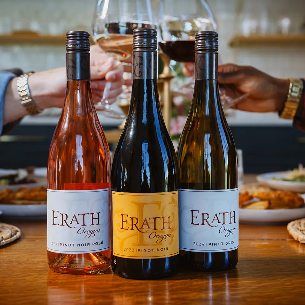 Three bottles of Erath wine on a table with people raising their wine glasses to toast in the background.